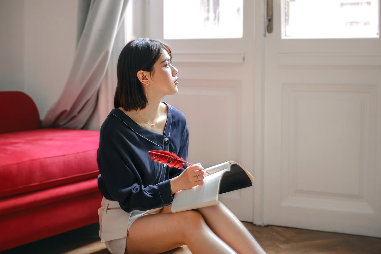 A young woman sits thoughtfully by a window, holding a feather pen and notebook, in a serene room.